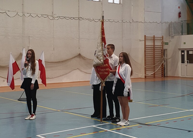Uczniowie w galowych strojach przy sztandarze szkoły na hali gimnastycznej.