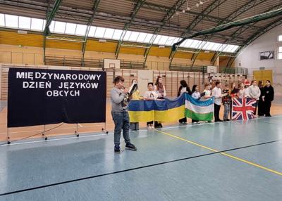 Uczniowie trzymający flagi różnych  narodowości  stojący na hali sportowej  i występujący przed zgromadzonymi uczniami i nauczycielami.