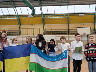 Uczniowie trzymający flagi różnych  narodowości  stojący na hali sportowej  i występujący przed zgromadzonymi uczniami i nauczycielami.