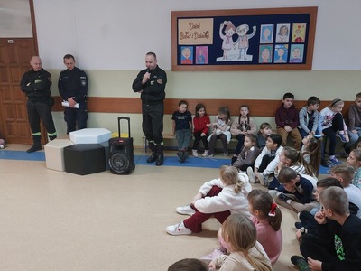 Strażacy i policjanci na korytarzu szkolnym pośród dzieci i młodzieży słuchających prelekcji.