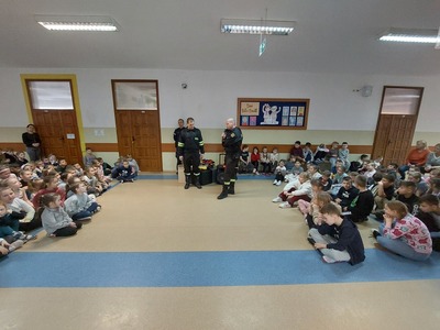 Strażacy i policjanci na korytarzu szkolnym pośród dzieci i młodzieży słuchających prelekcji.