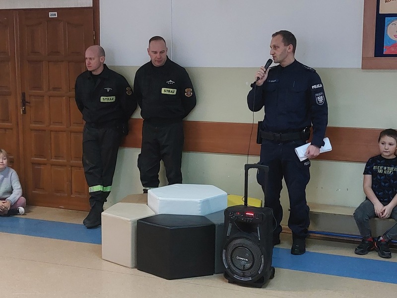 Strażacy i policjanci na korytarzu szkolnym pośród dzieci i młodzieży słuchających prelekcji.