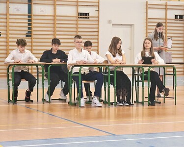 Uczniowie w strojach galowych na hali sportowej występujący przed społecznością szkolną.