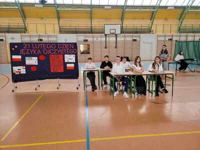 Uczniowie w strojach galowych na hali sportowej występujący przed społecznością szkolną.