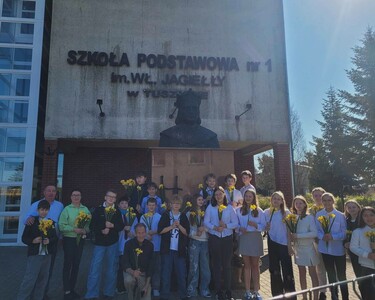 Uczniowie w raz z nauczycielem , dyrektorem szkoły przed budynkiem szkoły. przy pomniku patrona