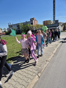 Uczniowie przemierzający ulice miast pod opieką nauczycieli trzymający zielone balony, chorągiewki oraz bibułowe kwiaty.