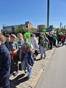 Uczniowie przemierzający ulice miast pod opieką nauczycieli trzymający zielone balony, chorągiewki oraz bibułowe kwiaty.
