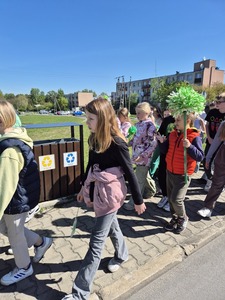 Uczniowie przemierzający ulice miast pod opieką nauczycieli trzymający zielone balony, chorągiewki oraz bibułowe kwiaty.