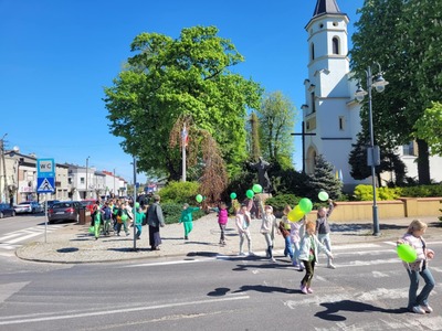 Uczniowie przemierzający ulice miast pod opieką nauczycieli trzymający zielone balony, chorągiewki oraz bibułowe kwiaty.