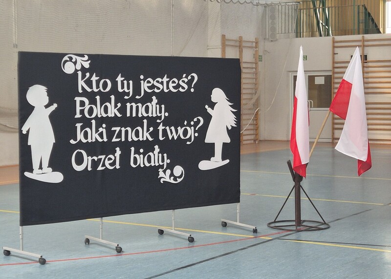Dekoracja do apelu przedstawiająca na grantowym tle postacie dziewczynki i chłopca a pomiędzy pierwsze dwa wersy  z wiersza. Obok dekoracji stoją flagi Polski.