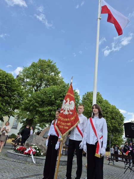 Uczniowie w strojach galowych ze sztandarem szkoły stojący na placu pod masztem z flagą biało-czerwoną.