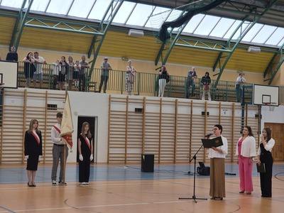 Hala sportowa . Poczet sztandarowy stojący na boku a przednim przemawiająca pani dyrektor w towarzystwie przewodniczącej szkoły oraz wicedyrektor.
