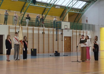 Hala sportowa . Poczet sztandarowy stojący na boku a przednim przemawiająca pani dyrektor w towarzystwie przewodniczącej szkoły oraz wicedyrektor.