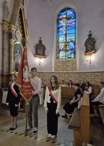 Poczet sztandarowy na mszy w kościele . Trójka uczniów. Dwie  dziewczyny ubrane w stroje galowe, między nim chłopak trzymający sztandar w stroju galowym.