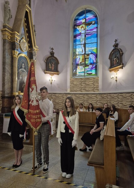 Poczet sztandarowy na mszy w kościele . Trójka uczniów. Dwie  dziewczyny ubrane w stroje galowe, między nim chłopak trzymający sztandar w stroju galowym.
