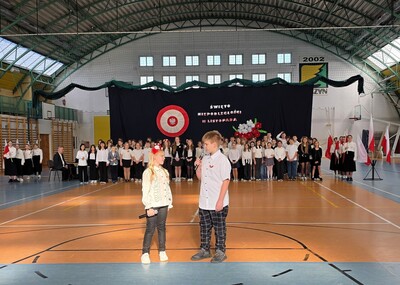 Uczniowie w strojach galowych  występujący przed zgromadzoną społecznością szkolną. na hali sportowej.