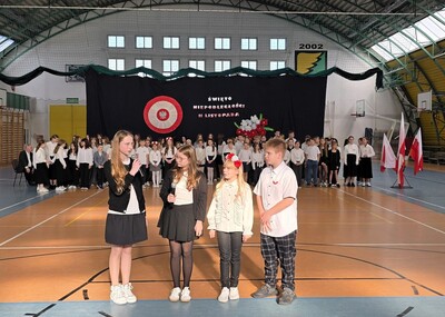 Uczniowie w strojach galowych  występujący przed zgromadzoną społecznością szkolną. na hali sportowej.