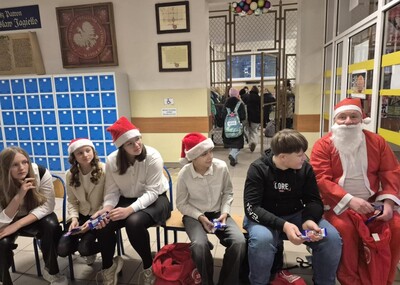 Uczniowie wraz ze Świętym Mikołajem witający uczniów w holu szkoły na wejściu  i obdarowując ich słodkim upominkiem.