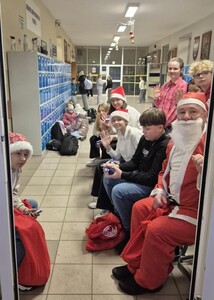 Uczniowie wraz ze Świętym Mikołajem witający uczniów w holu szkoły na wejściu  i obdarowując ich słodkim upominkiem.