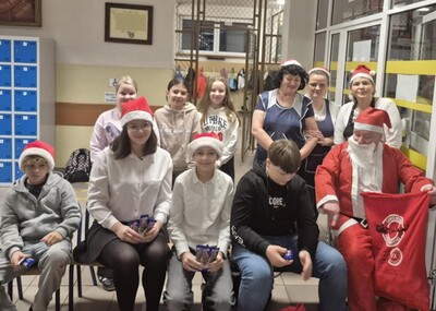 Uczniowie wraz ze Świętym Mikołajem witający uczniów w holu szkoły na wejściu  i obdarowując ich słodkim upominkiem.