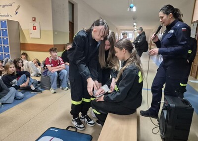 Uczniowie  w strojach strażackich na holu demonstrujący zgromadzonym sposób udzielania pierwszej pomocy.
