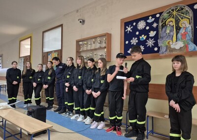 Uczniowie  w strojach strażackich na holu szkoły występujący przed zgromadzonymi uczestnikami apelu.