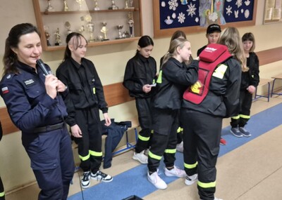 Uczniowie  w strojach strażackich wraz z przedstawicielem policji na holu szkoły występujący przed zgromadzonymi uczestnikami apelu.