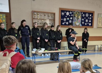 Uczniowie  w strojach strażackich na holu szkoły występujący przed zgromadzonymi uczestnikami apelu.