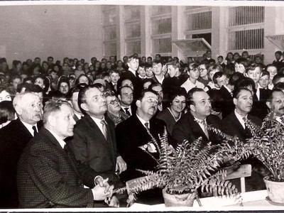 Ludzie zgromadzeni na sali gimnastycznej. Dwa kwiatki w doniczkach postawione na stole. W tle po prawej stronie okna z metalowymi kratami.