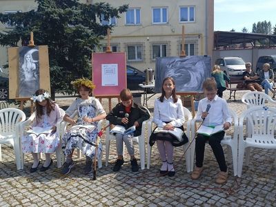 Pięcioro uczniów klas piątych, w tym dwie uczennice w strojach świtezianek, siedzi na krzesłach i czytają fragmenty Ballad i romansów Mickiewicza.
