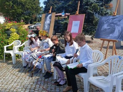 Pięcioro uczniów, w tym dwie uczennice w strojach świtezianek, siedzi na krzesłach i czytają fragmenty Ballad i romansów Mickiewicza.