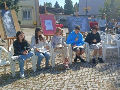 Pięcioro uczniów siedzi na krzesłach i czyta fragmenty Ballad i romansów Mickiewicza. Za nimi na sztalugach okładka książki i obrazy lokalnego malarza.