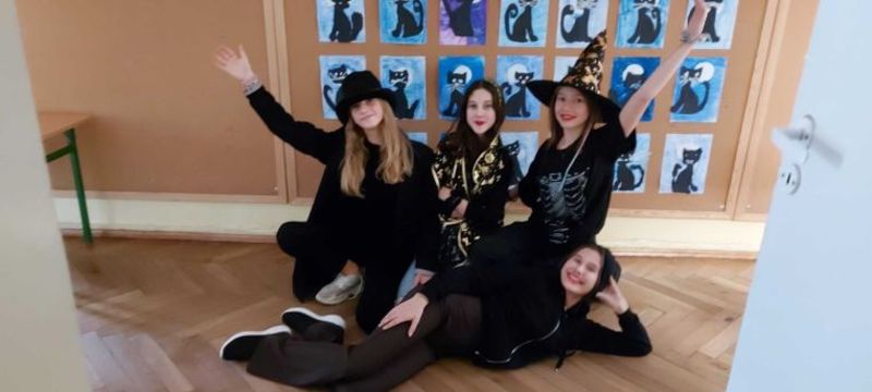 Cztery uczennice w strojach wróżek. Wszystkie ubrane na czarno, na głowach kapelusze. Trzy kucają, jedna leży na boku na podłodze, głowę podpiera ręką.