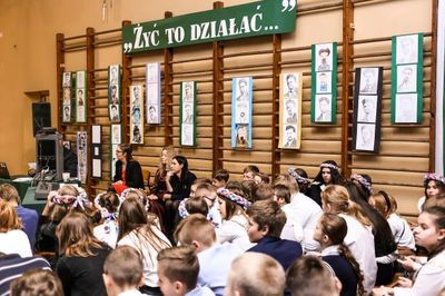 Widownia. Na niskich ławkach siedzą uczniowie. W tle drabinki z zawieszonymi na nich portretami Reymonta.
