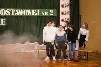 Scena na sali gimnastycznej. Obok ławki grupa stojących uczniów ubranych w typowe uczniowskie stroje. Widoczny dym unoszący się nad podłogą.