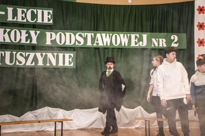 W centralnej części aktor wcielający się w rolę Reymonta. Ubrany w czarny frak, na jego głowie cylinder.