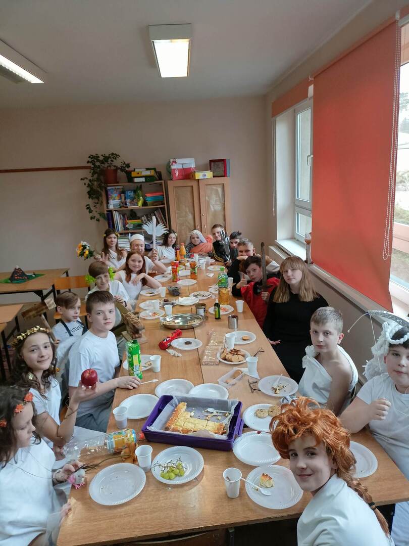 Uczniowie klasy 5 b zgromadzeni w sali lekcyjnej przy długim stole zastawionym smakołykami. Dzieci przebrane w stroje bogów i bogiń greckich.