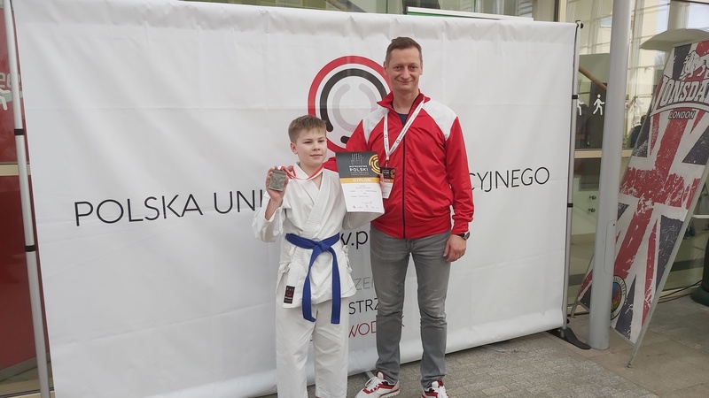 Kacper wraz ze stojącym obok trenerem. Chłopiec ubrany w strój karateki trzyma w jednej ręce medal, w drugiej dyplom. 