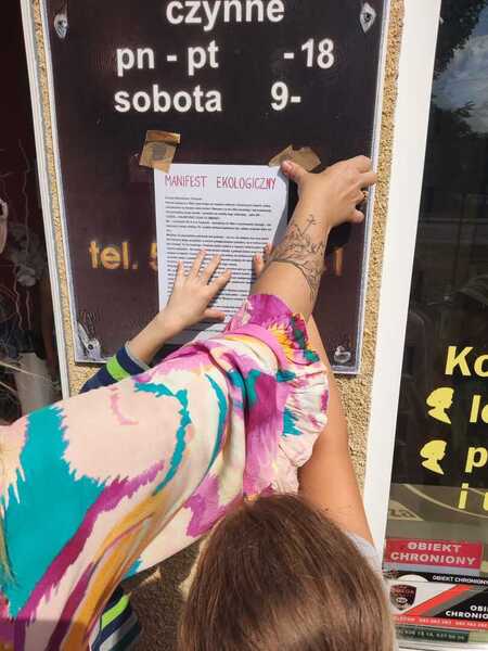 Pracownica lokalu wieszająca manifest ekologiczny obok godzin otwarcia obiektu.