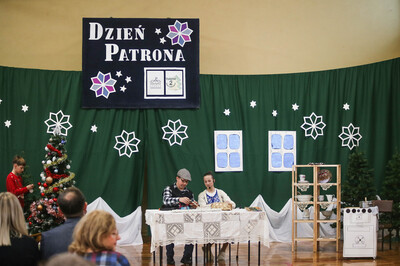 Sala gimnastyczna. Sceneria zimowa. Na środku stół, przy nim siedzi dwoje uczniów - aktorów. Po bokach choinki. Na górze napis: DZIEŃ PATRONA