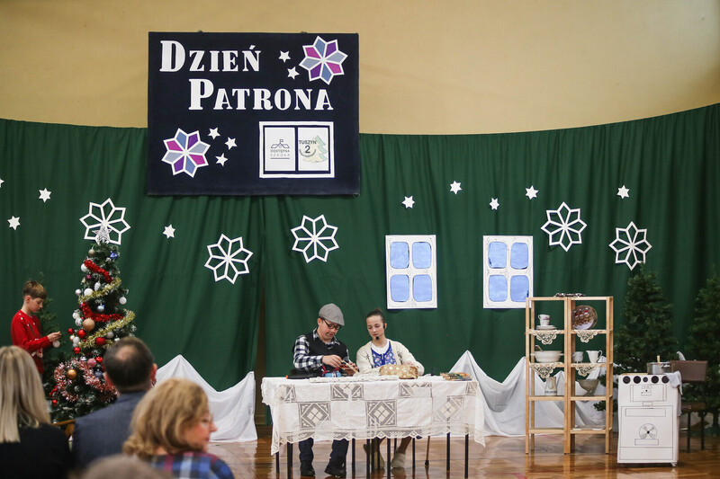 Sala gimnastyczna. Sceneria zimowa. Na środku stół, przy nim siedzi dwoje uczniów - aktorów. Po bokach choinki. Na górze napis: DZIEŃ PATRONA