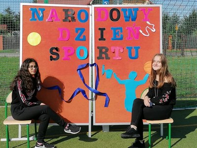 Na zdjęciu uczennice na tle kolorowego napisu MIĘDZYNARODOWY DZIEŃ SPORTU.