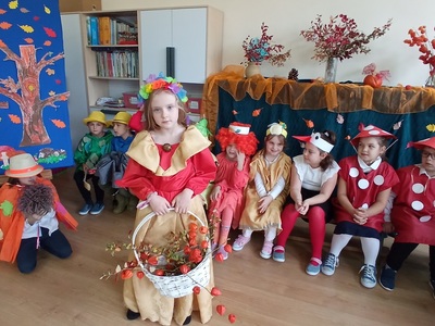 Dziewczynka z koszem w ręku, z tyłu grupa uczniów na tle jesiennej dekoracji.