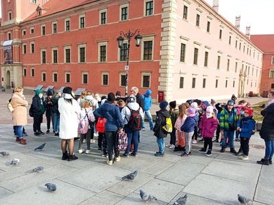 Grupa uczniów z opiekunami i przewodnikiem na Placu Zamkowym w Warszawie.