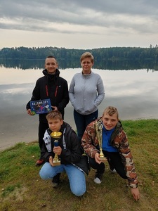 Cztery osoby stoją z pucharami i nagrodami. W tle jezioro.