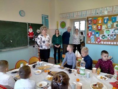 Dzieci siedzą przy stole z tyłu stoi pani dyrektor i pracownicy szkoły składają życzenia..