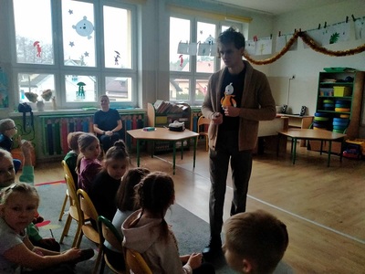Dzieci siedzą na krzesełkach przed nimi stoi pan.