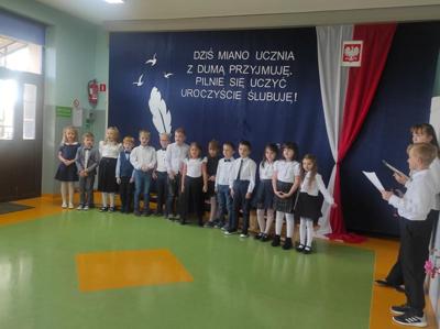 Uczniowie stoją w jednym rzędzie na tle dekoracji. Z prawej strony widać osoby prowadzące uroczystoś