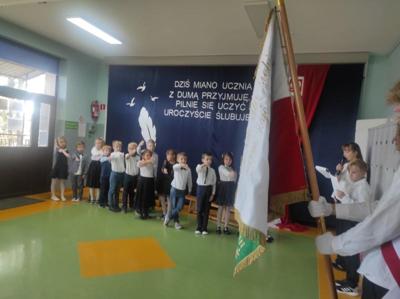 Uczniowie z podniesioną prawą ręką skierowani są w stronę sztandaru szkoły.