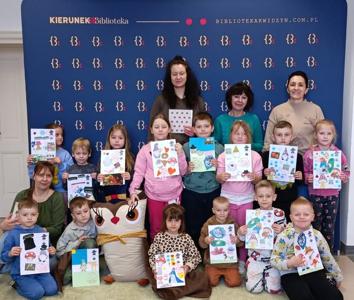 Zdjęcie przedstawia przedszkolaki, wychowawcę i opiekuna wycieczki wraz z pracownikami biblioteki. Na zdjęciu dzieci trzymają wykonane podczas warsztatów prace. 